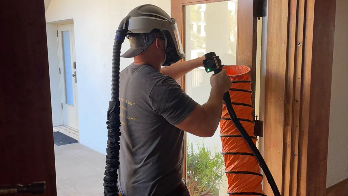 Green Bee Laser technician in a powered respirator laser cleaning a wood-framed glass door, with a fume extraction hose positioned at the work area